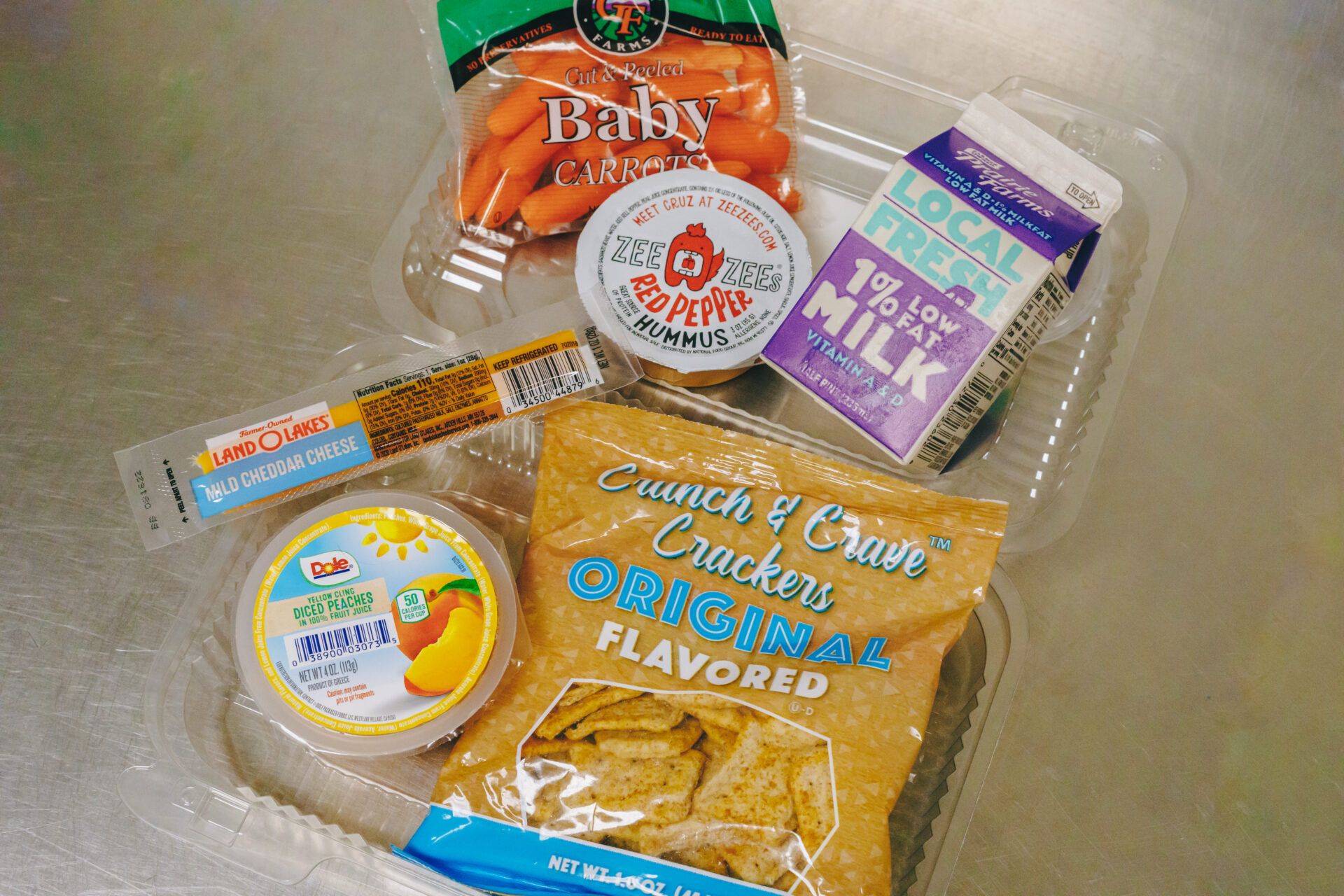 Image of a to-go school meal with milk, crackers, hummus, cheese, carrots and peaches.