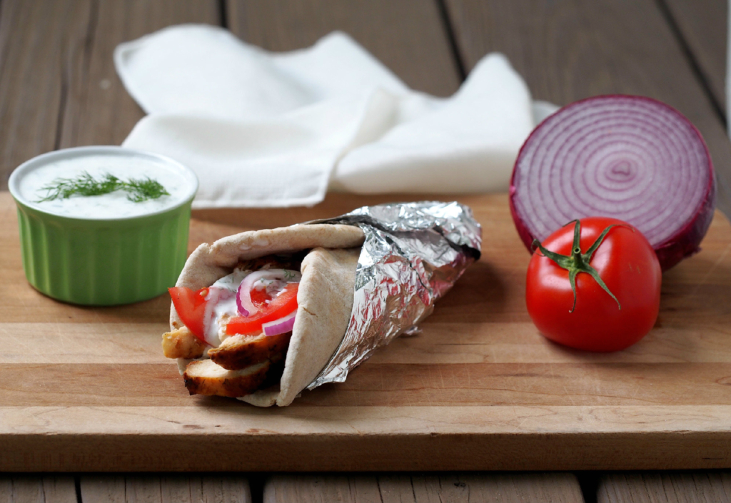 Grilled Chicken Gyros with Tzatkiki Sauce on a cutting board next to a red onion and tomato.
