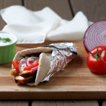 Grilled Chicken Gyros with Tzatkiki Sauce on a cutting board next to a red onion and tomato.