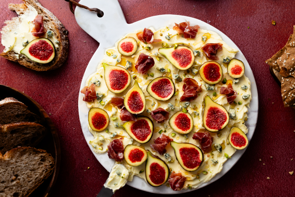 Overhead image of a butter board topped with sliced figs, prosciutto, bleu cheese and nuts.