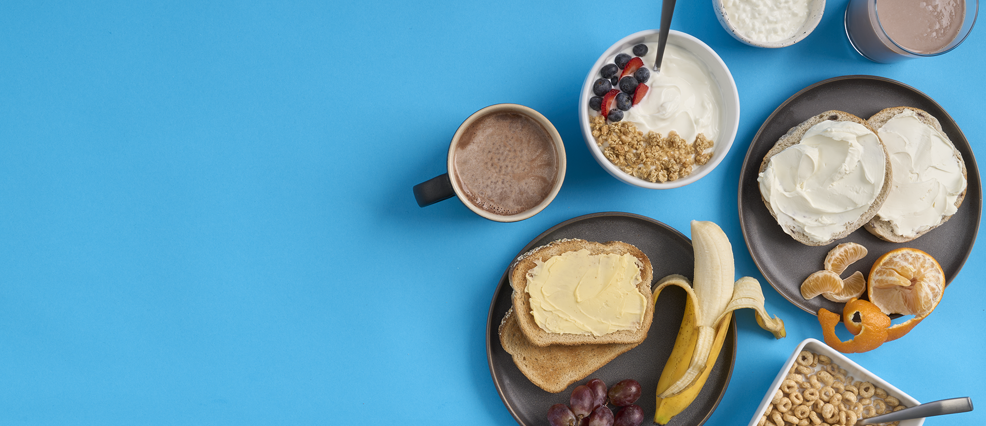 A array of breakfast foods on plates and in bowls, hot chocolate, parfait, bagel and cream cheese, butter toast, banana, cereal, cottage cheese, chocolate milk, and an orange on a bright blue background.