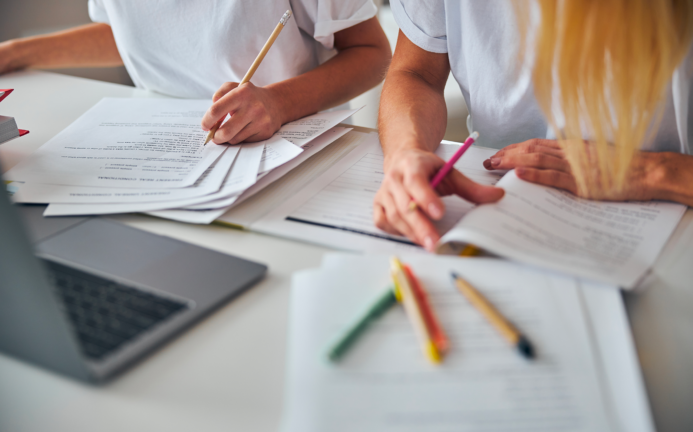 Two young professionals look at a computer and stacks of paper while working.