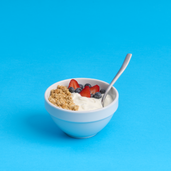 Bowl of vanilla yogurt with granola and berries with spoon on a bright blue background.