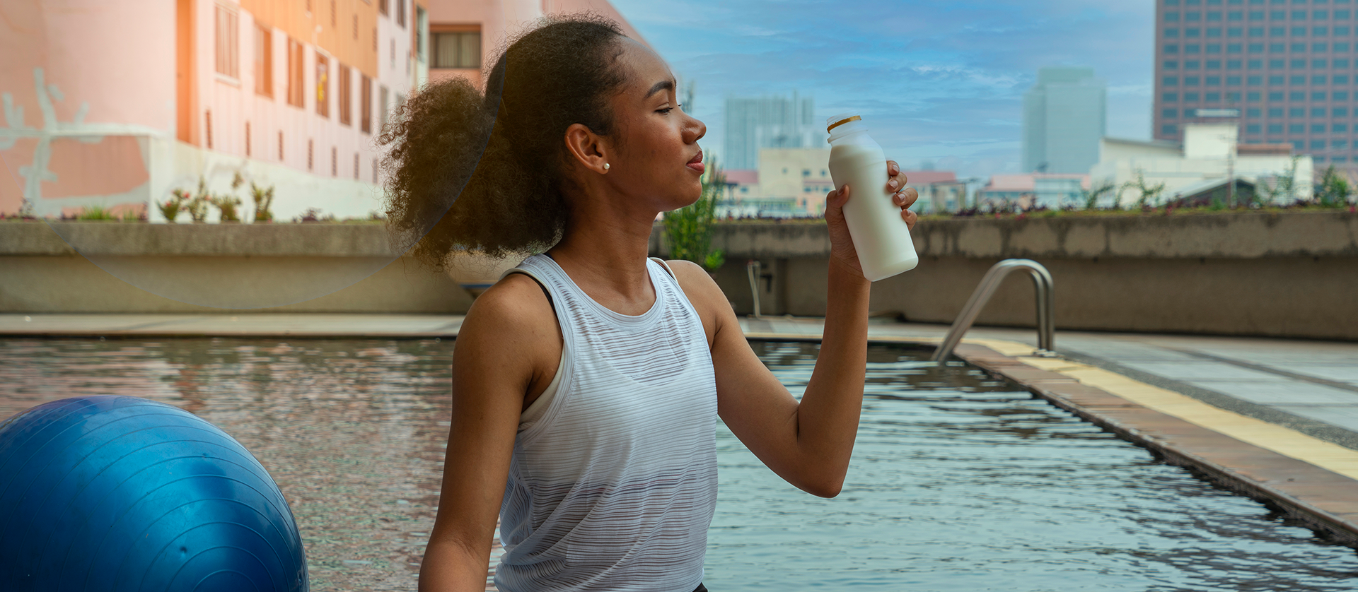 Woman exercising outside in front of a swimming pool in a white tank top drinking white milk out of a milk chug.