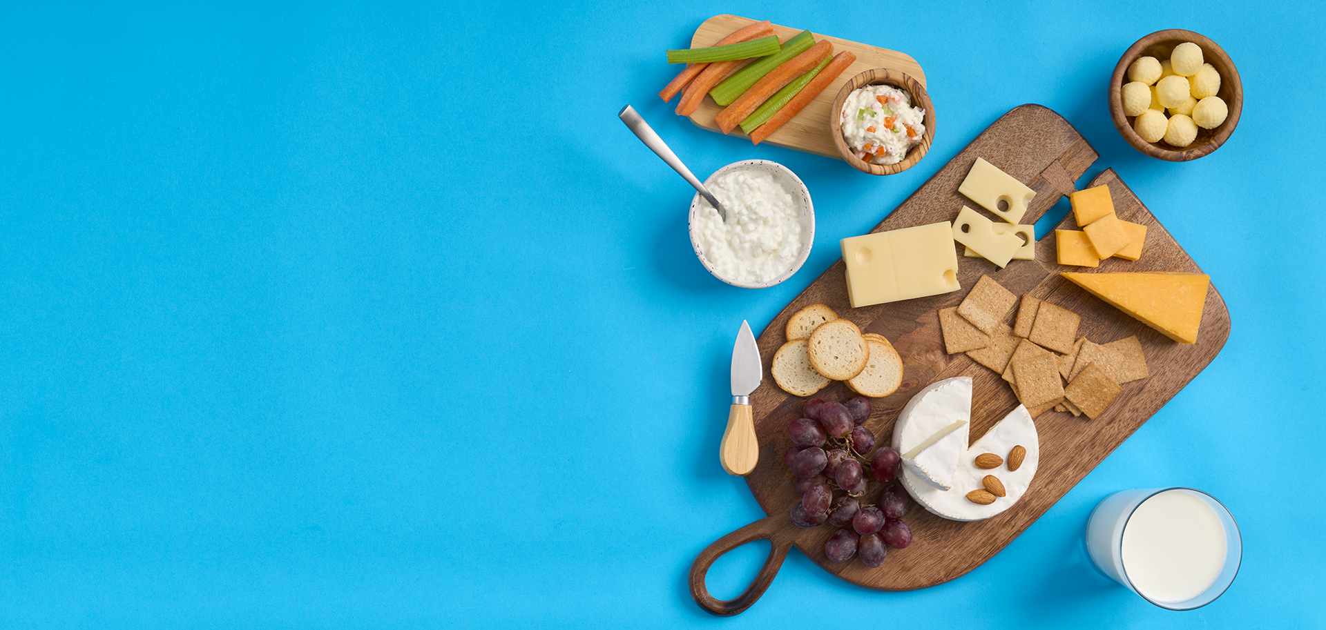 Tray of cheese and crackers, butter, cottage cheese, veggie dip and celery sticks with carrots, and a glass of white milk on a bright blue background