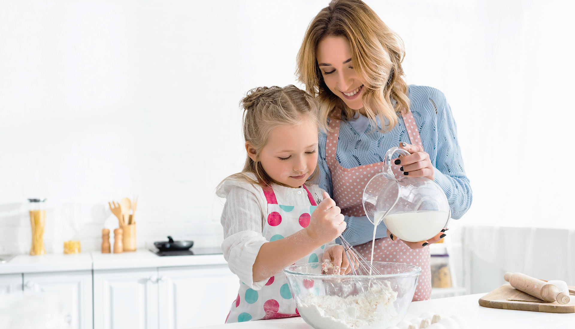 Daughter kneading dough while mother pouring milk from jug in bowl at home.