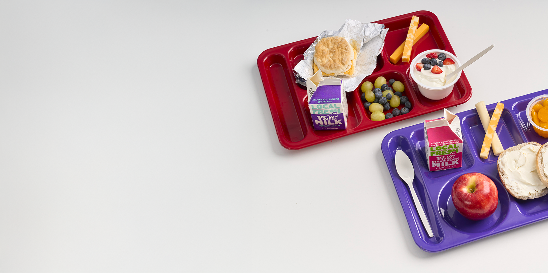 Two trays of school breakfasts with fruit and cheese sticks and a carton of white milk and stawberry milk on a white table.