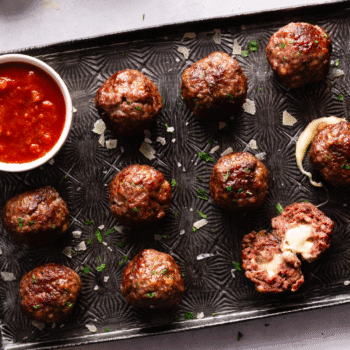 Image of mozzarella stuffed meatballs on a baking sheet with a small bowl of marinara sauce to the side.